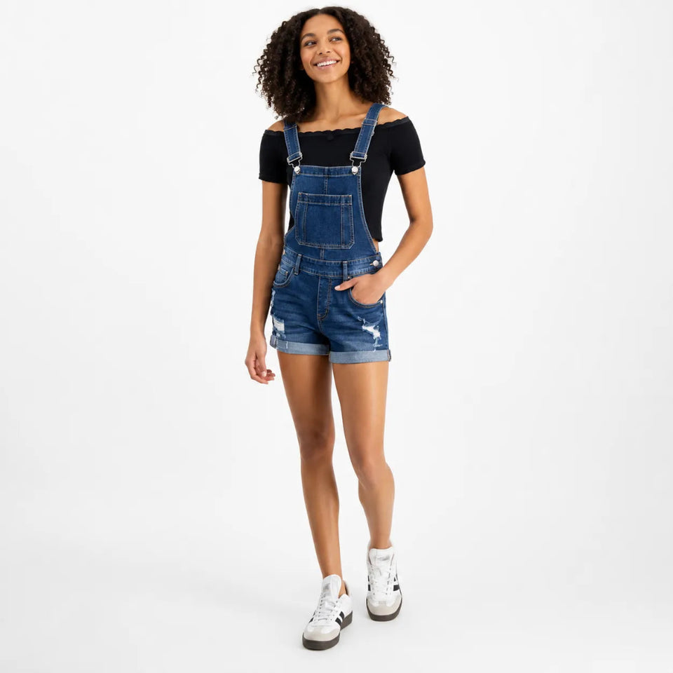 A woman modeling a pair of blue denim overall shorts with distressed details, worn over a black off-the-shoulder t-shirt and paired with white sneakers, against a plain white studio background.