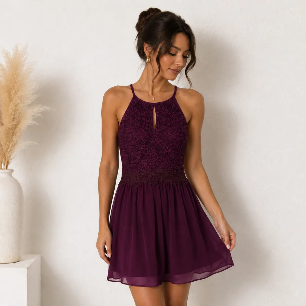 A woman modeling a sleeveless halter mini dress in deep burgundy purple. The dress features a lace-embroidered bodice with a keyhole cutout and a flowing chiffon skirt, set against a neutral white wall with a decorative vase of pampas grass on the left.
