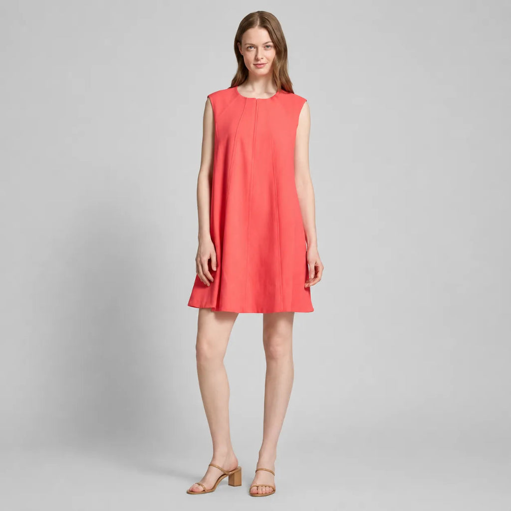 A woman modeling a sleeveless coral pink shift dress with a round neckline and a flared silhouette, paired with tan heeled sandals, against a light gray studio background.