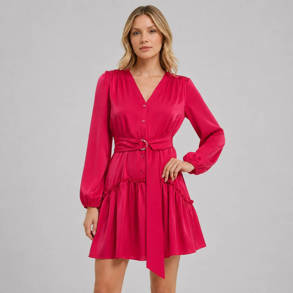 A woman modeling a bright pink long-sleeve mini dress with a V-neck, button front, and matching tie belt, photographed against a plain light gray studio background.