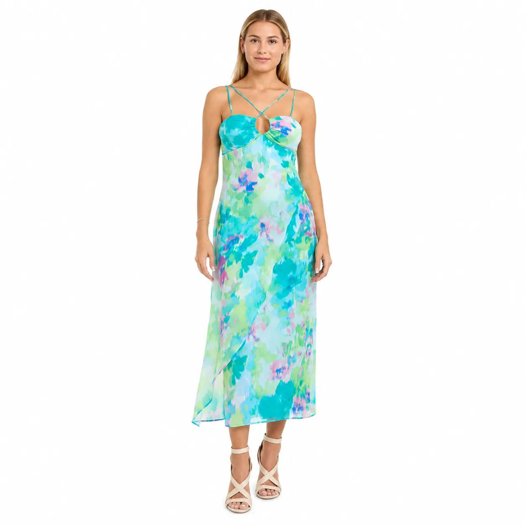 A woman modeling a sleeveless midi dress with thin straps and a watercolor floral print in shades of turquoise, green, and purple, paired with beige strappy high-heeled sandals against a plain white background.