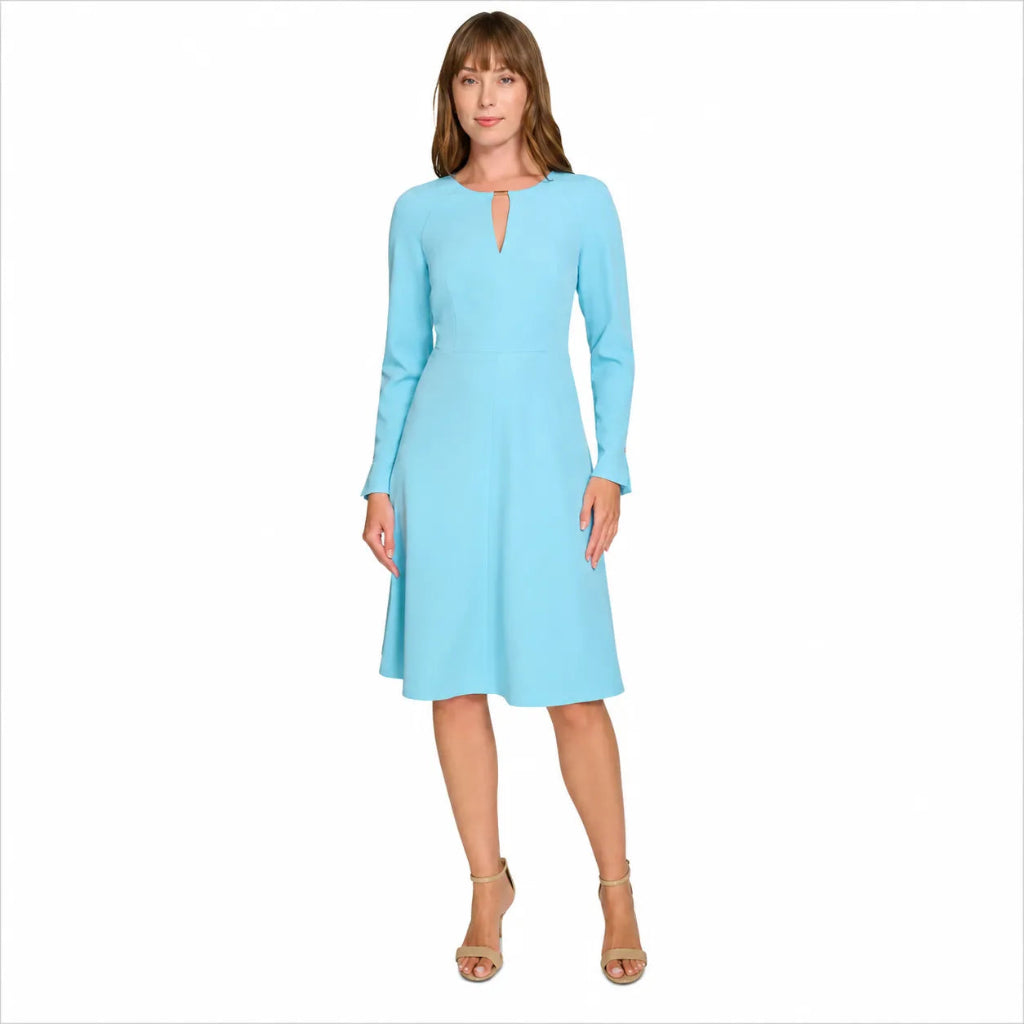 A woman modeling a light blue long-sleeve knee-length dress with a small keyhole cutout at the neckline, paired with beige high-heeled sandals, against a plain white background.
