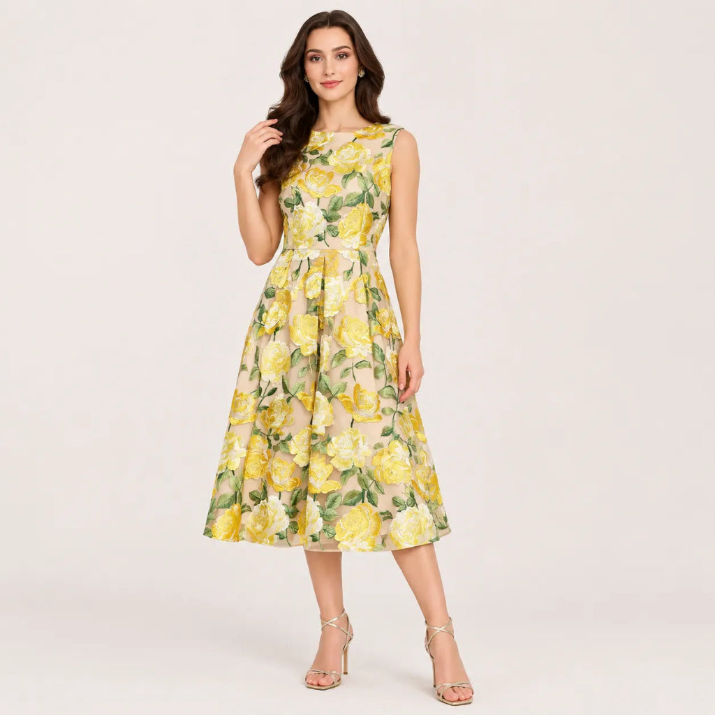 A woman modeling a sleeveless, knee-length A-line dress with a yellow and green floral print on a light beige background, paired with clear high-heeled sandals, against a solid light gray studio backdrop.
