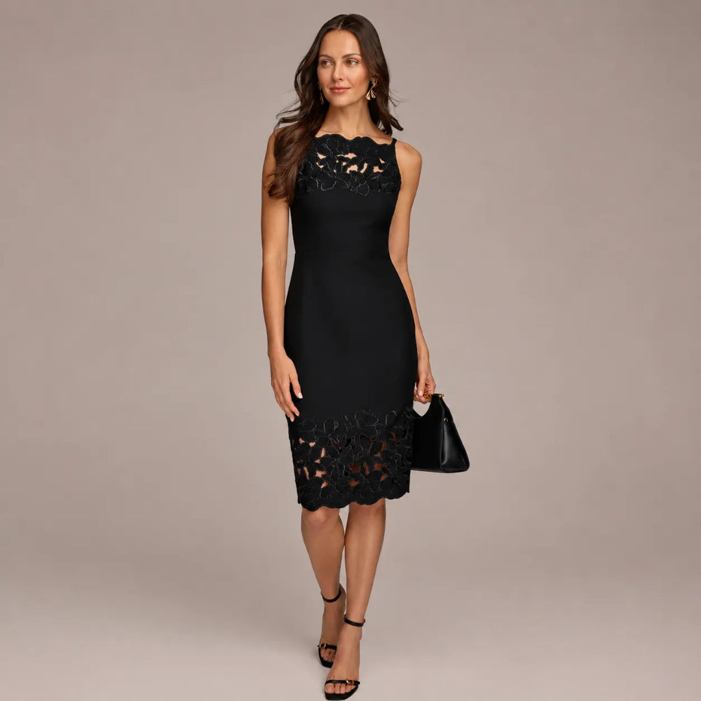 A woman models a sleeveless black lace dress with a fitted silhouette and scalloped lace details at the neckline and hem, paired with black high-heeled sandals and a small black handbag, against a solid warm gray studio background.