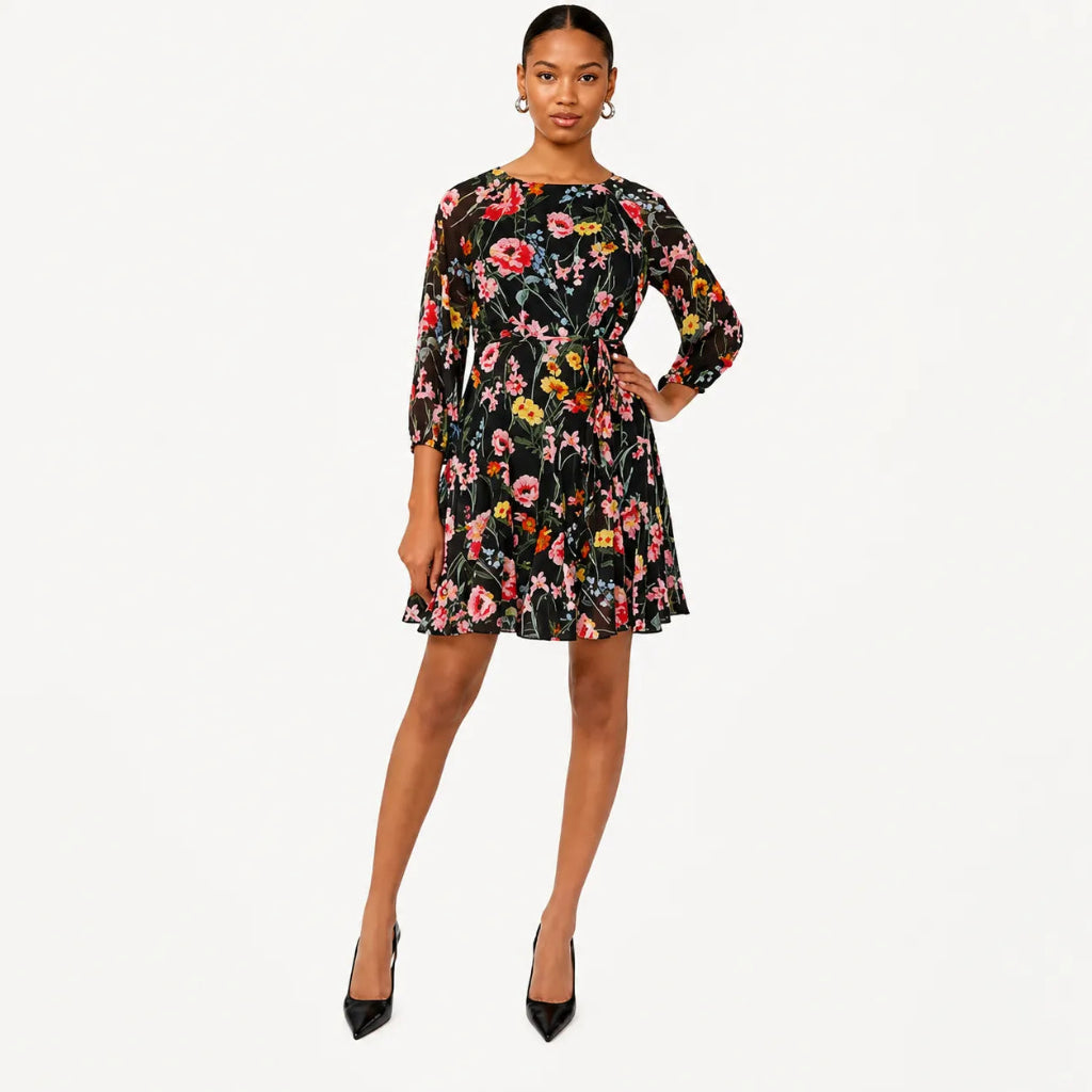 A woman modeling a short black floral dress with three-quarter sleeves and a flared skirt, paired with black pointed heels, against a light gray studio background.