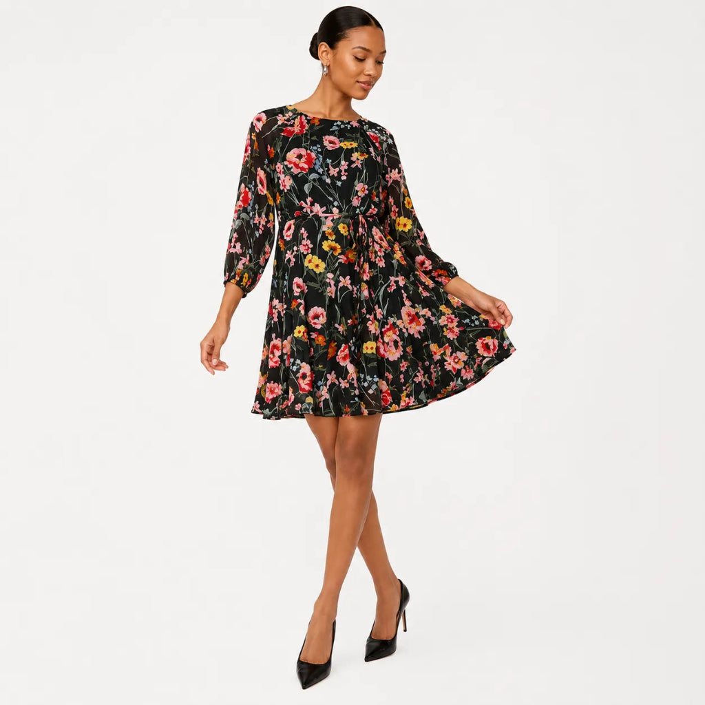 A woman modeling a black three-quarter sleeve mini dress with a vibrant pink, red, and yellow floral print, paired with black pointed-toe heels, against a plain white studio background.