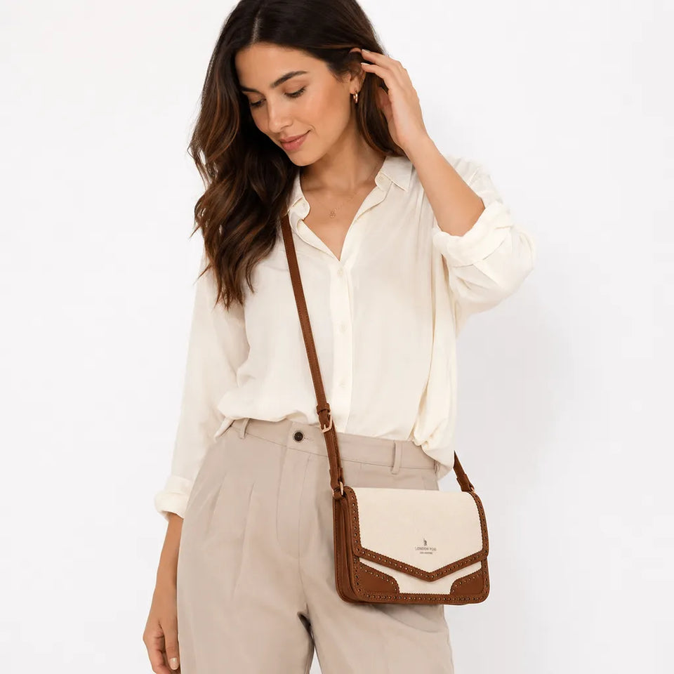 A woman wearing a cream button-up shirt and beige trousers carries a small crossbody bag with a cream and brown color-blocked design and a brown strap, set against a plain white studio background.