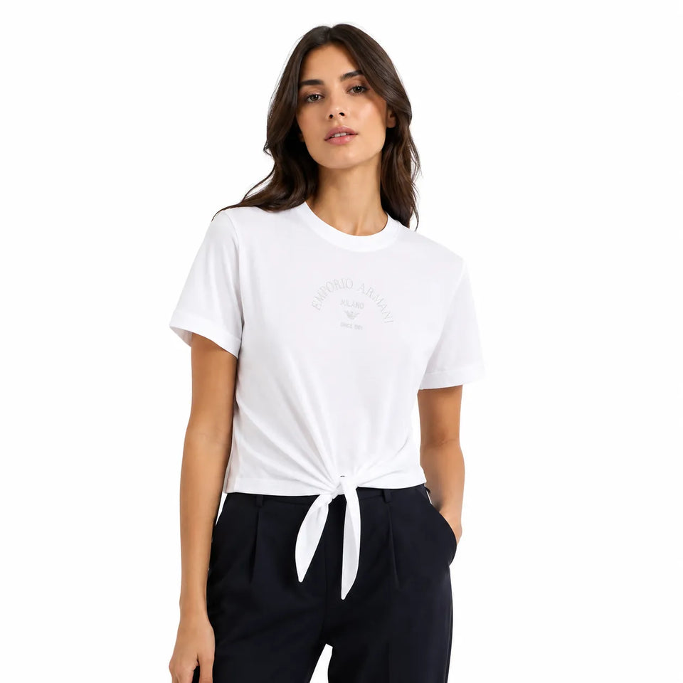 A woman wearing a white short-sleeve crewneck t-shirt with a subtle tonal graphic on the chest, styled with dark trousers and tied at the waist, against a plain white background.