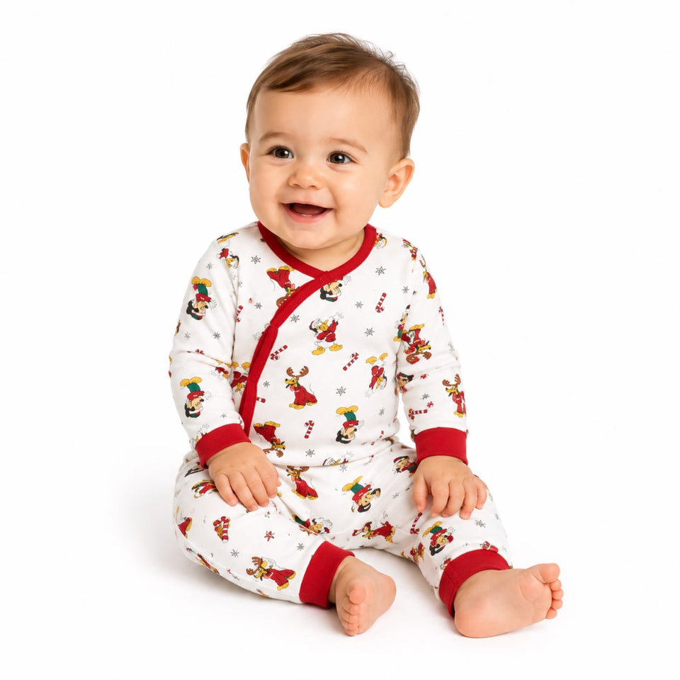 A smiling baby sitting on a white studio background wearing a white one-piece pajama with red trim and a festive holiday print.