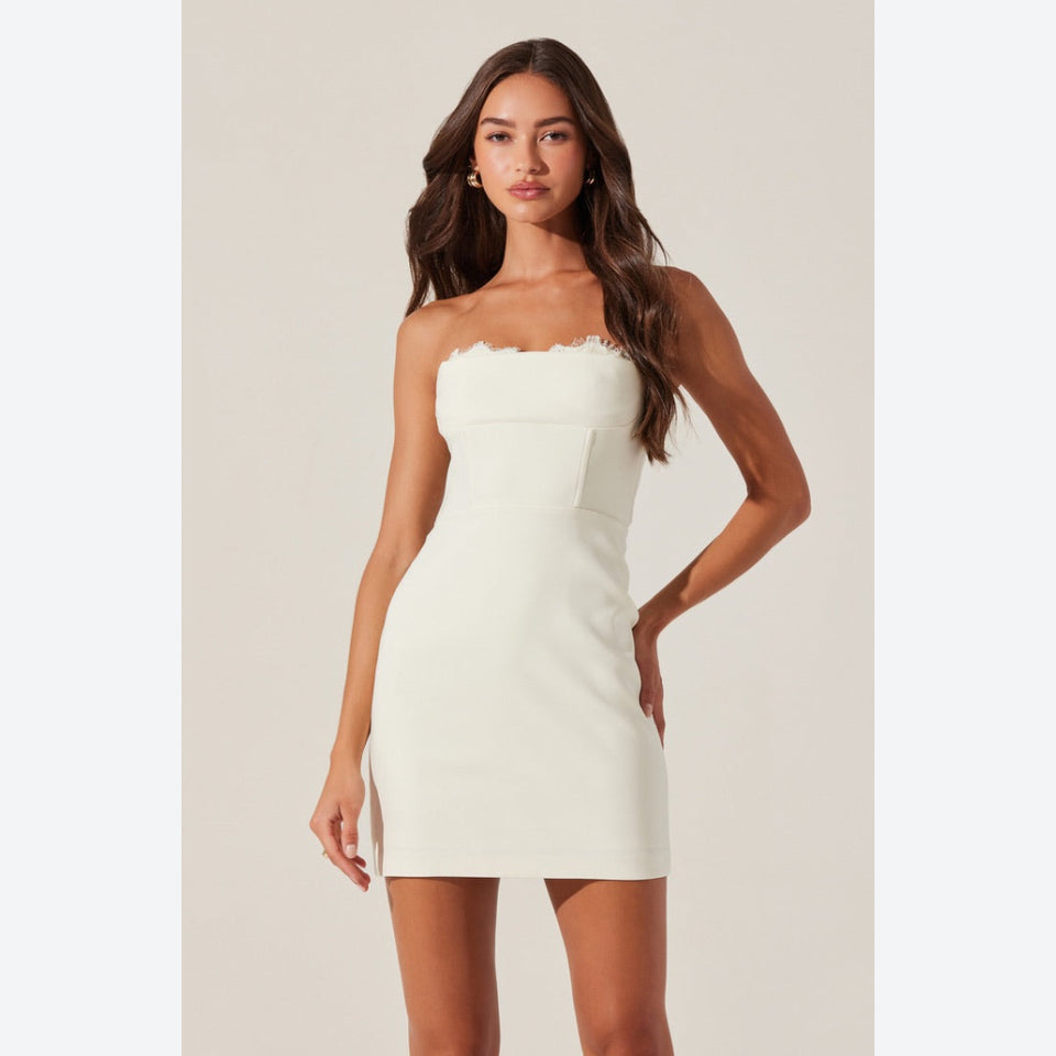 A woman modeling a strapless off-white bodycon mini dress with a scalloped neckline, photographed against a plain light beige studio background.