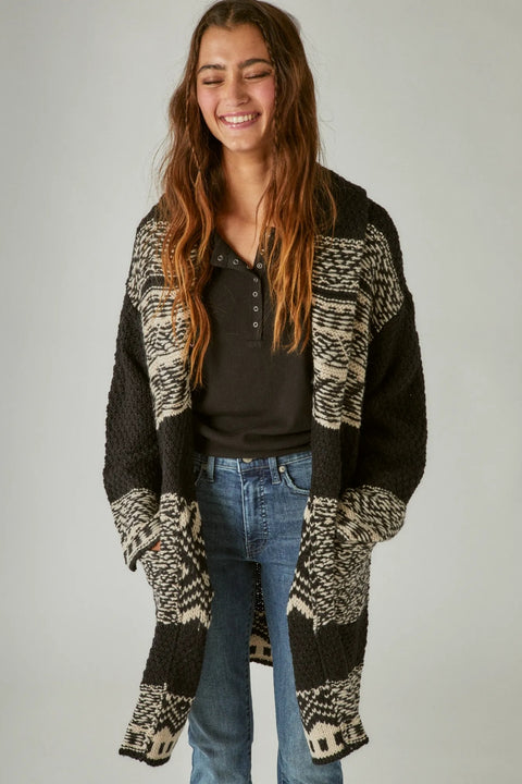 Woman wearing a patterned black and white cardigan over a dark shirt and jeans on a plain background