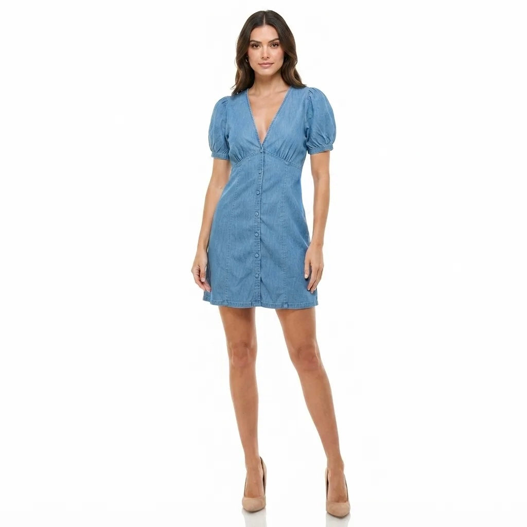 A woman modeling a light blue chambray mini dress with puff sleeves, a V-neckline, and a button-up front, set against a plain white background.