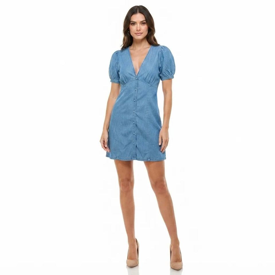 A woman modeling a light blue chambray mini dress with puff sleeves, a V-neckline, and a button-up front, set against a plain white background.