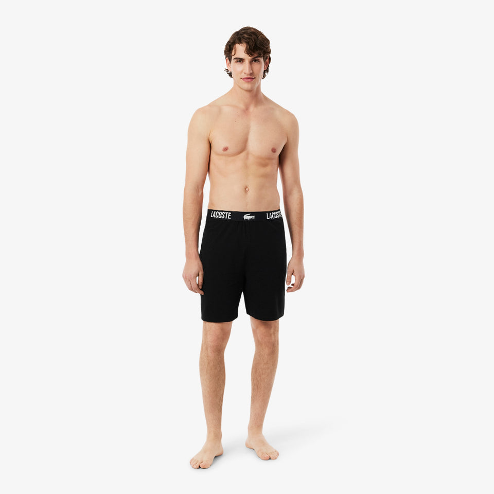 A male model stands against a light gray studio background wearing black boxer briefs with a white Lacoste logo band.