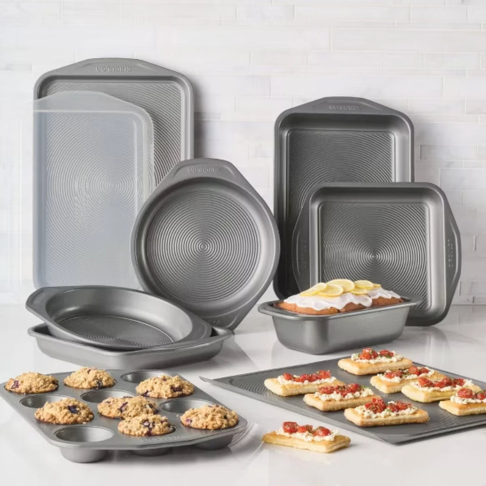 A 15-piece non-stick baking pan set in dark gray, displayed on a white countertop against a white subway tile backsplash. The set includes various rectangular, round, and square pans, a muffin tin, and a pizza pan, with some baked goods (muffins, cookies, pizza) shown in use.