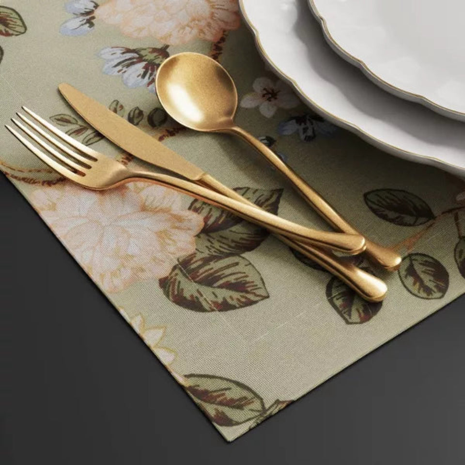 Gold-colored flatware, including a fork, knife, and spoon, arranged on a beige floral placemat next to white plates.