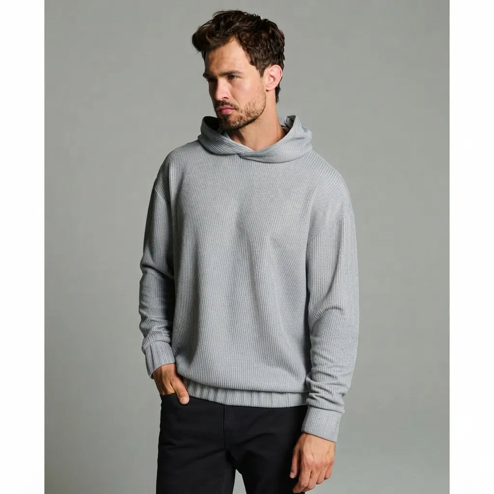 A man wearing a light gray ribbed knit hooded sweater and dark pants, standing against a solid gray studio background.