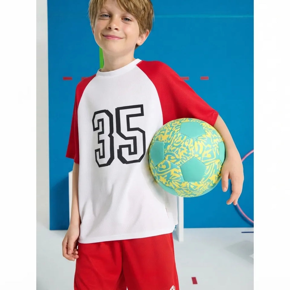 A young boy wearing a white and red raglan t-shirt with the number 35 on the front, red shorts, and holding a green and yellow soccer ball against a blue studio background.