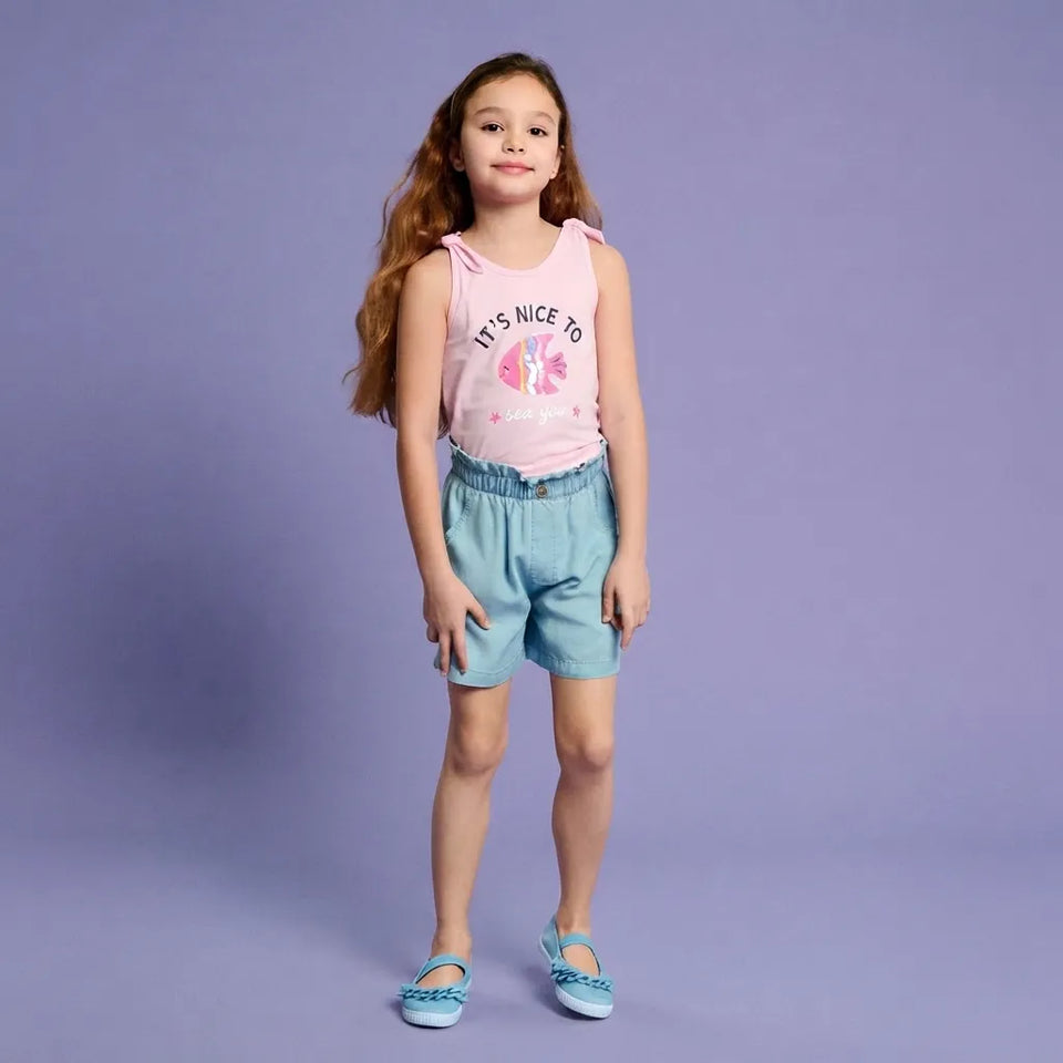 A young girl stands against a solid lavender studio background wearing a light pink sleeveless tank top with a peace sign graphic and the text 'IT'S NICE TO', light blue denim shorts, and light blue flat shoes.