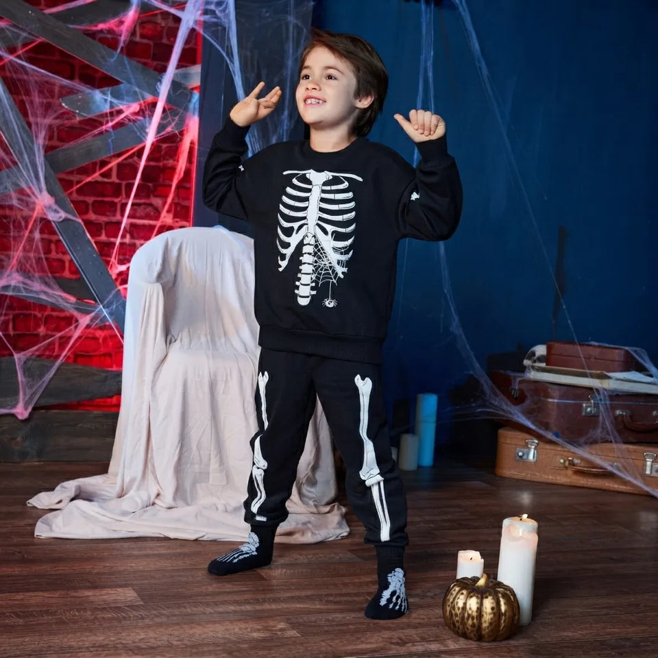 A young boy modeling a black Halloween sweatshirt and matching sweatpants set with white skeleton graphics, posing in a spooky themed room with cobwebs, candles, and a ghosted chair.