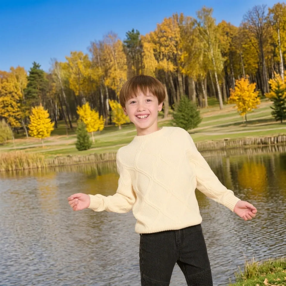 A young boy smiling and posing outdoors by a lake, wearing a cream-colored cable-knit sweater and dark pants, with a background of autumn trees and blue sky.