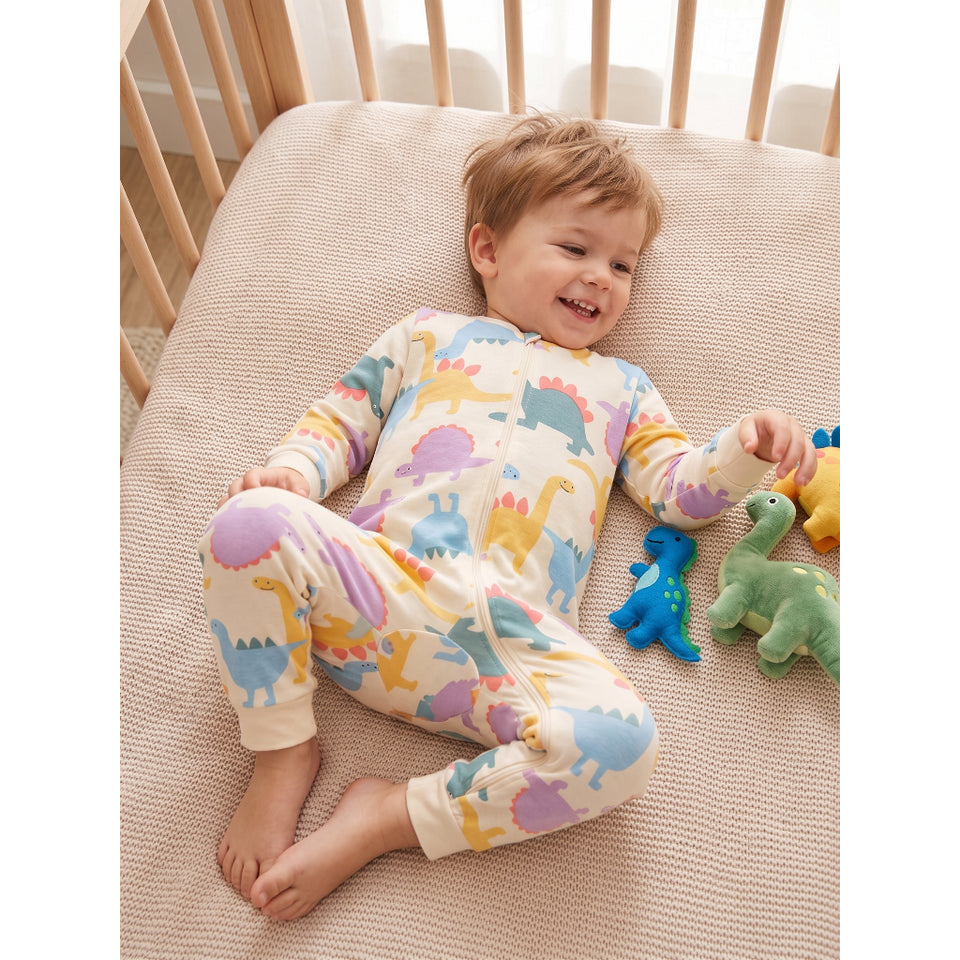 A baby lying on a textured beige mattress in a wooden crib, wearing a cream-colored long-sleeve footless pajama romper covered in colorful dinosaur illustrations. The baby is smiling and interacting with plush dinosaur toys.