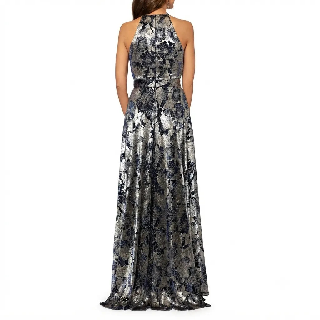 A sleeveless halter-neck floor-length gown with a metallic silver and navy blue abstract floral pattern, shown from the back on a model against a white background.