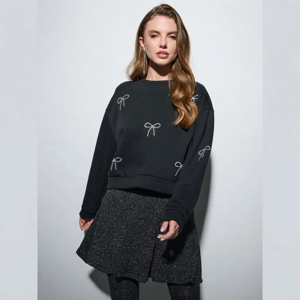 A woman modeling a black crewneck sweatshirt decorated with white embroidered bow motifs, paired with a black textured skirt, against a light gray studio background.