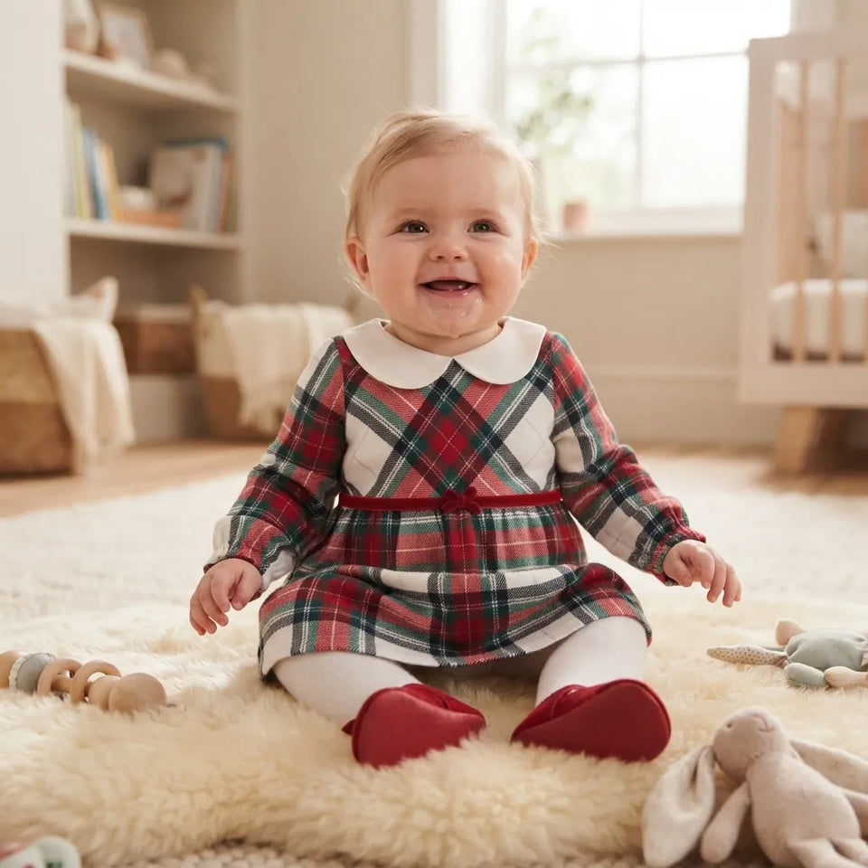 A baby sitting on a fluffy cream rug in a bright nursery, wearing a red, green, and white tartan plaid dress with a white Peter Pan collar and red booties.