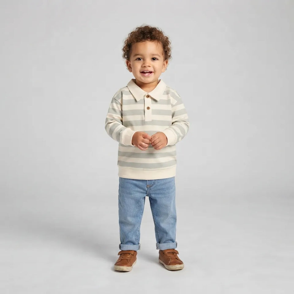 A toddler standing against a light gray studio background, wearing a cream and light gray striped long-sleeve polo shirt, light blue jeans, and brown shoes.