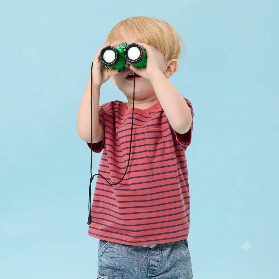 A young child with blond hair wearing a red and navy striped t-shirt and blue jeans, holding a green toy binocular up to their eyes against a solid light blue background.