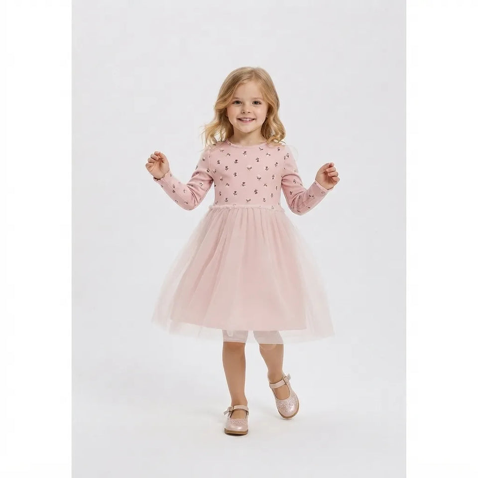 A young girl with blonde hair stands against a light gray studio background wearing a light pink long-sleeve dress with a tulle skirt, white tights, and pink shoes.