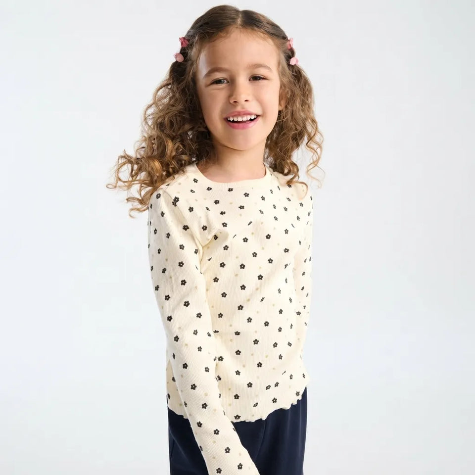 A young girl with curly brown hair wearing a cream-colored long-sleeve top with a black floral pattern, paired with navy blue pants, photographed against a solid light gray background.