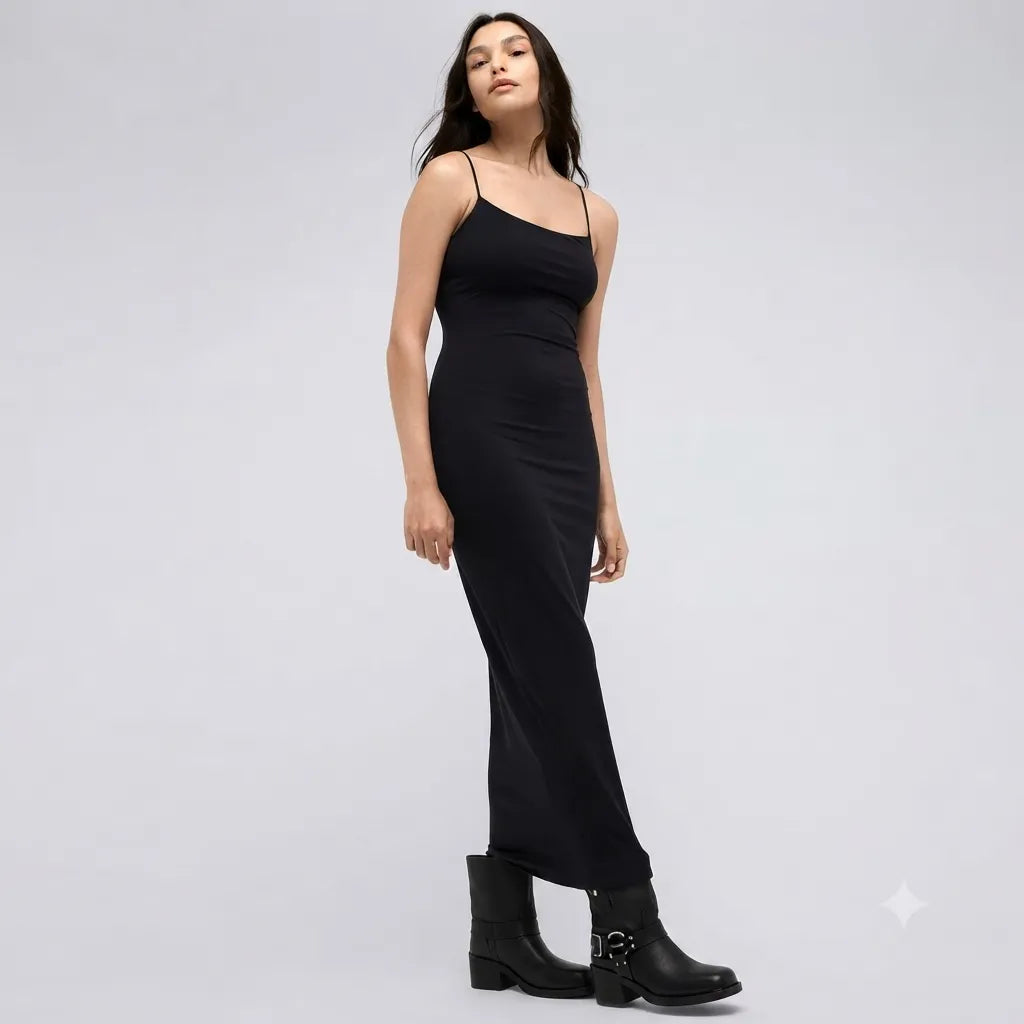 A woman modeling a black spaghetti-strap maxi dress with a fitted silhouette, paired with black ankle boots, against a light gray studio background.