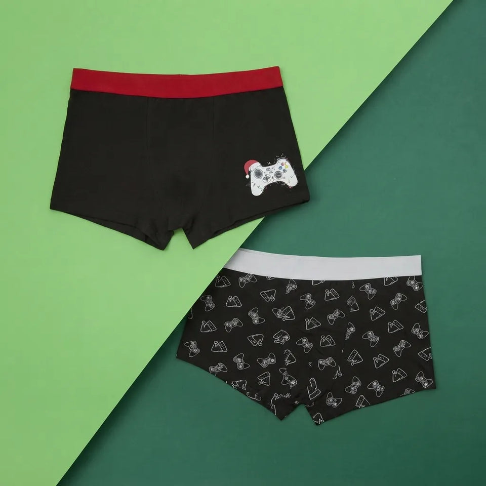 Two pairs of men's boxer briefs laid flat on a split green background. The top pair is black with a red waistband and a small white video game controller graphic with a red hat on the right side.