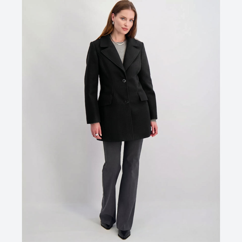 A woman modeling a black double-breasted coat with notched lapels, paired with dark gray trousers and black shoes, against a light gray studio background.