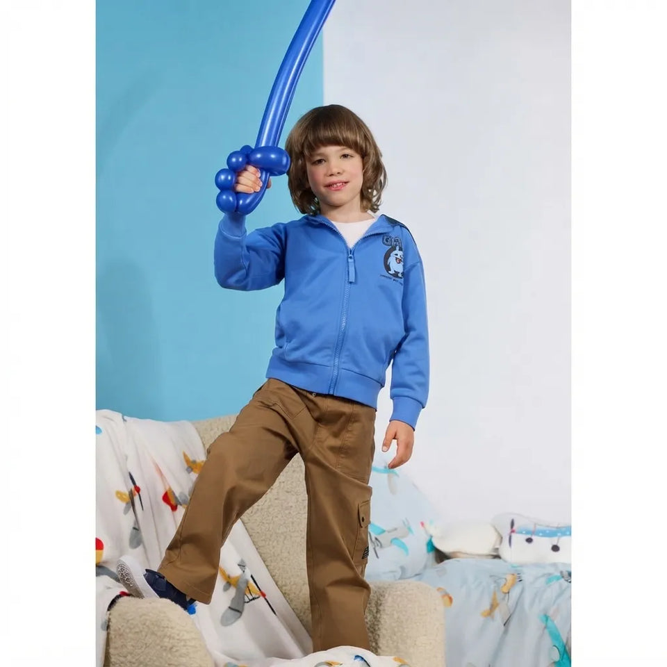 A young boy wearing a blue zip-up hoodie and brown pants stands on a beige chair in a bedroom setting, holding a blue balloon sword.