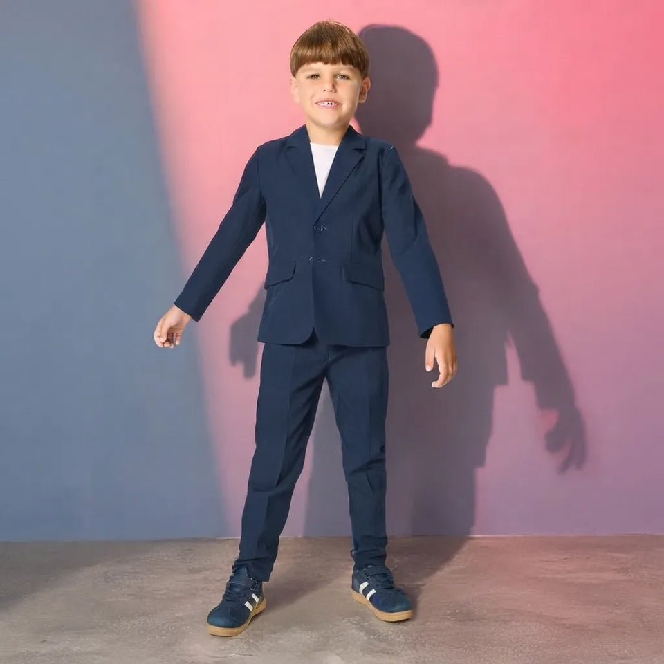 A young boy modeling a navy blue two-piece suit with a white t-shirt and navy sneakers, standing against a pink and blue gradient studio backdrop.
