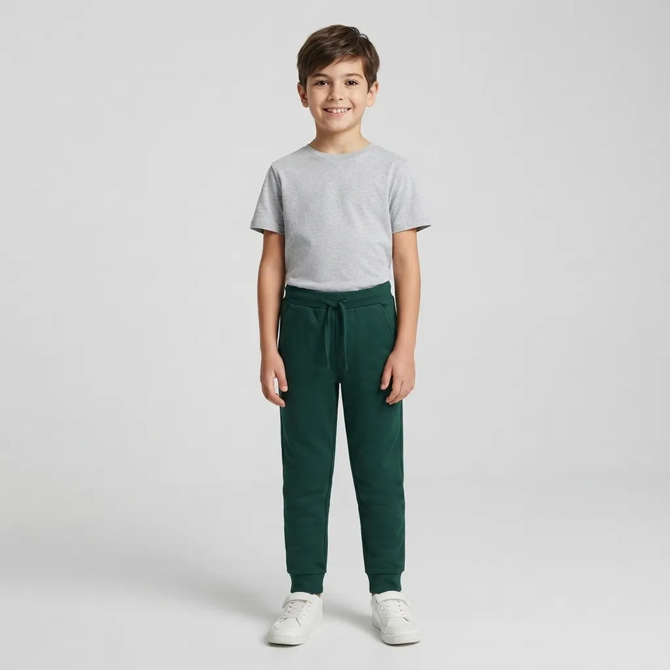 A young boy stands against a light gray studio background wearing a light gray crewneck t-shirt, dark green jogger-style pants, and white sneakers.