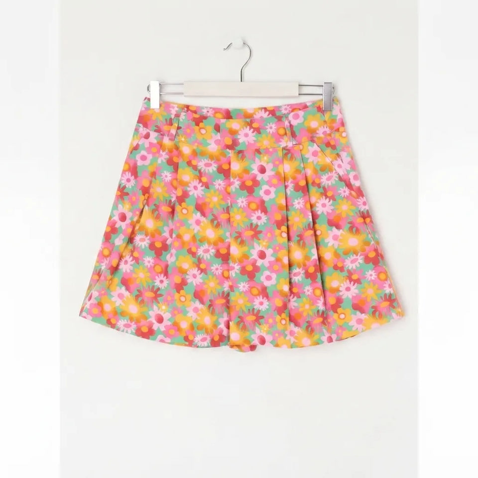 A short pleated skirt with a vibrant multicolor floral print, hanging on a white hanger against a plain white wall.