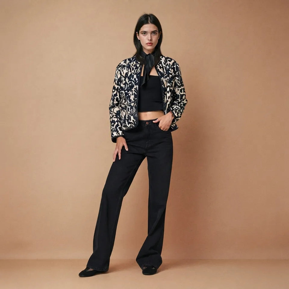 A model stands against a solid beige studio backdrop wearing a black and white abstract patterned jacket over a black crop top and black wide-leg trousers.