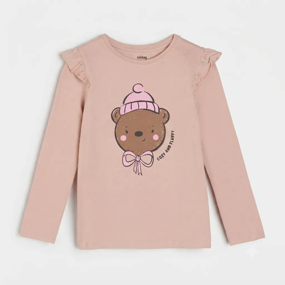 A light pink long-sleeve t-shirt for girls, featuring ruffled shoulders and a central graphic of a brown cartoon bear wearing a pink beanie and bow tie, with the text 'COD. AND CLASITY' printed next to it.