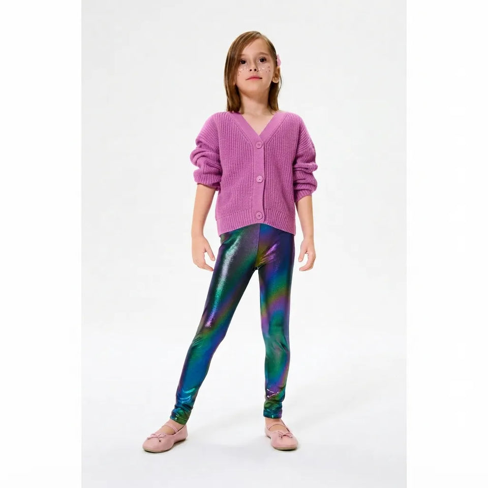 A young girl modeling shiny, iridescent leggings with a purple cardigan and pink shoes, standing against a plain white studio background.