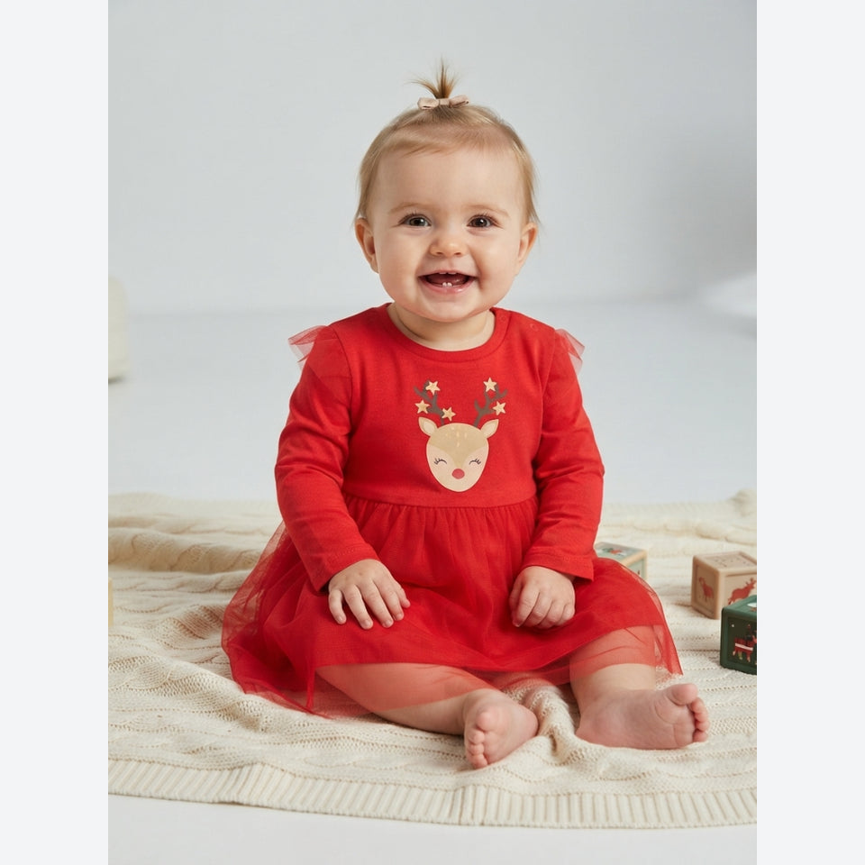 A baby girl sitting on a cream knit blanket wearing a festive red long-sleeve dress with a tulle skirt and a reindeer face applique on the chest.