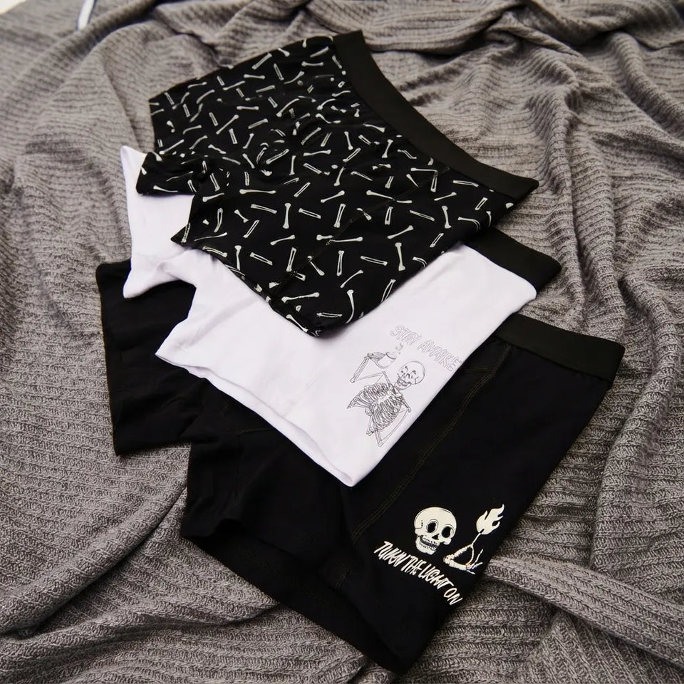 Three pairs of folded boxer briefs laid flat on a heather gray fabric background. One pair is black with a white graphic print, one is white with a black graphic print, and one is black with a white repeating pattern.