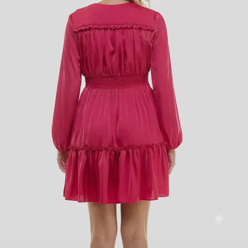 A woman wearing a bright pink long-sleeve mini dress with ruffled trim and a smocked waist, shown from the back against a light gray background.