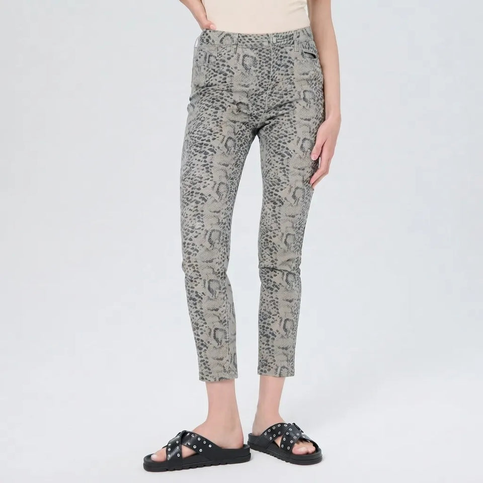 A model wearing slim-fit ankle-length pants with a gray and black snakeskin pattern, paired with black slide sandals and a beige top, shown against a plain light gray studio background.