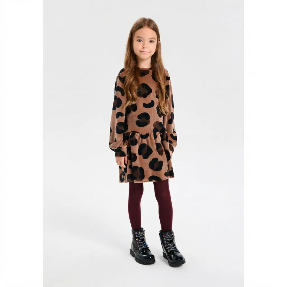 A young girl stands against a light gray studio background wearing a brown and black leopard-print long-sleeve dress, burgundy tights, and black lace-up boots.