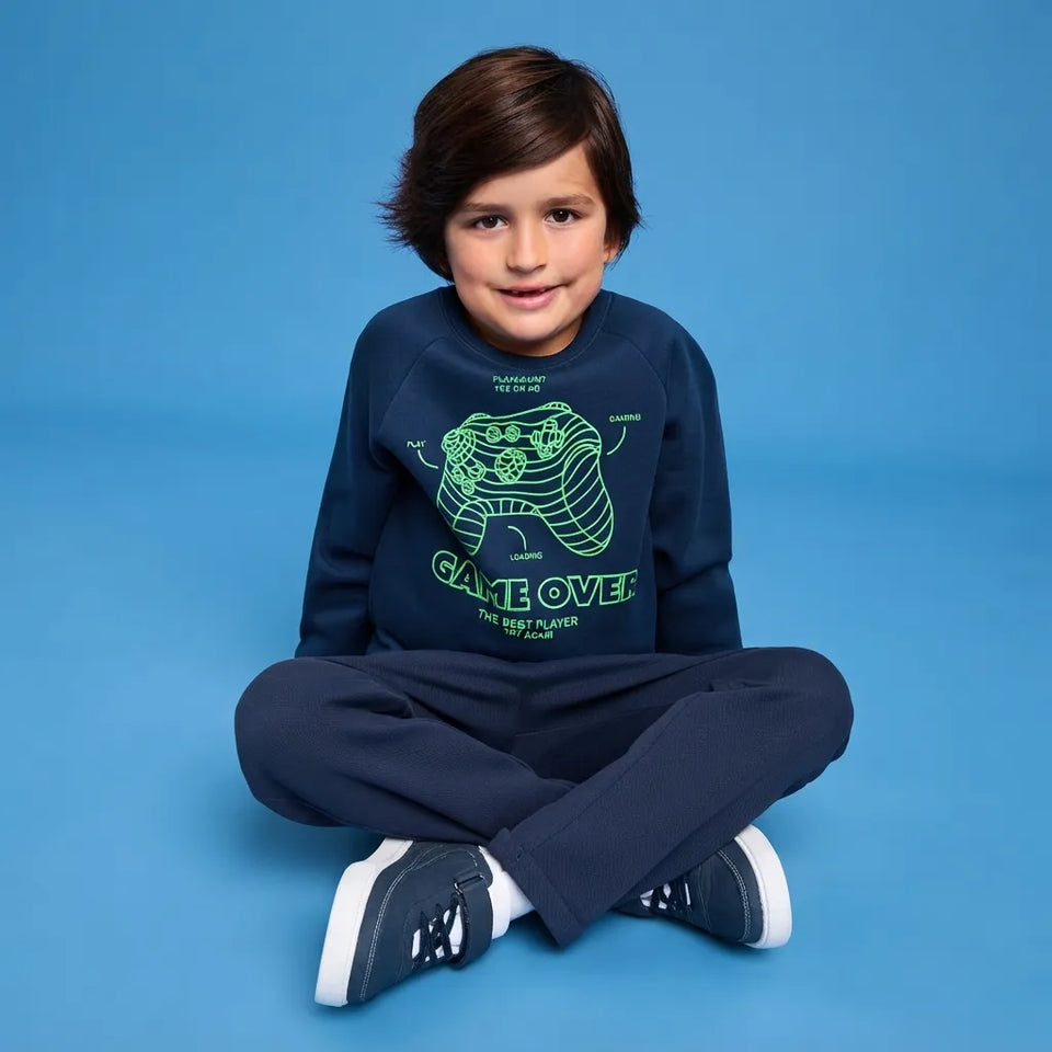 A young boy sits cross-legged on a solid blue studio background, wearing a navy blue sweatshirt with a green video game controller graphic and the text 'GAME OVER THE LOST PLAYER' paired with navy pants and sneakers.