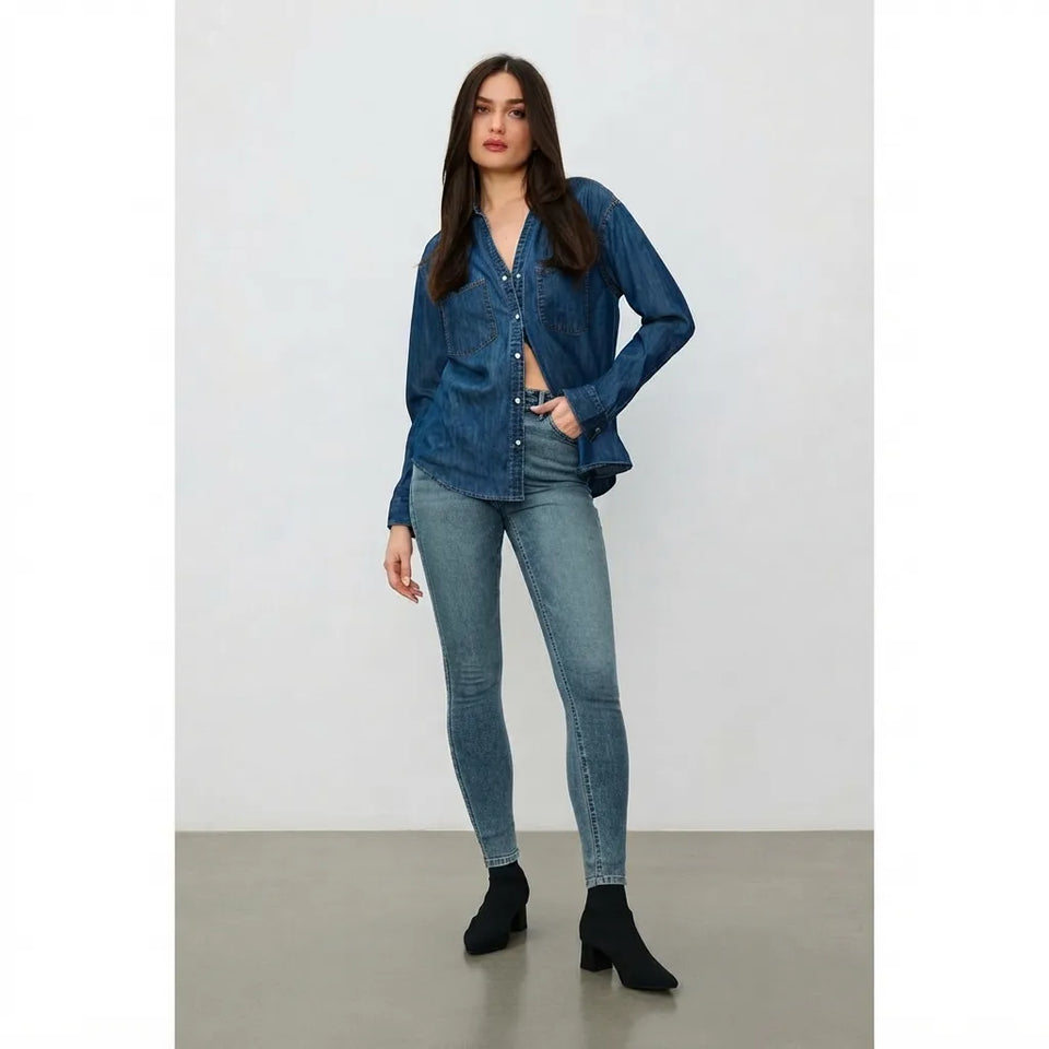 A woman modeling a dark blue denim button-up shirt and light blue skinny jeans with black ankle boots, standing against a neutral studio backdrop.